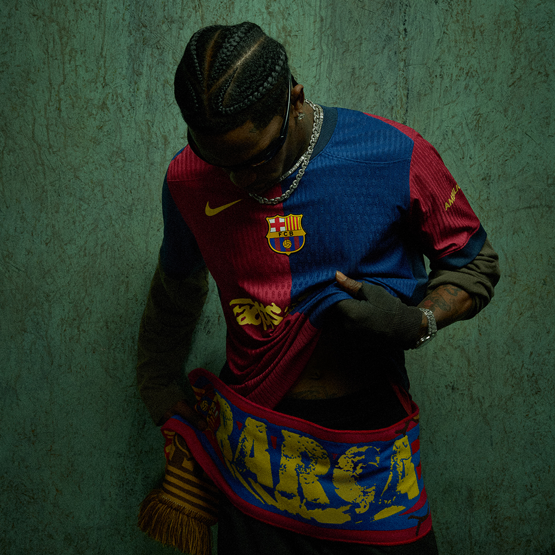 Travis Scott in a FC Barcelona jersey and scarf, looking down, with braided hair and sunglasses, in a dark, textured setting.